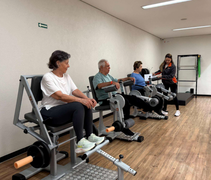 Pessoas idosas em equipamentos de fortalecimento de membros inferiores sob supervisão profissional em Academia Terapêutica.