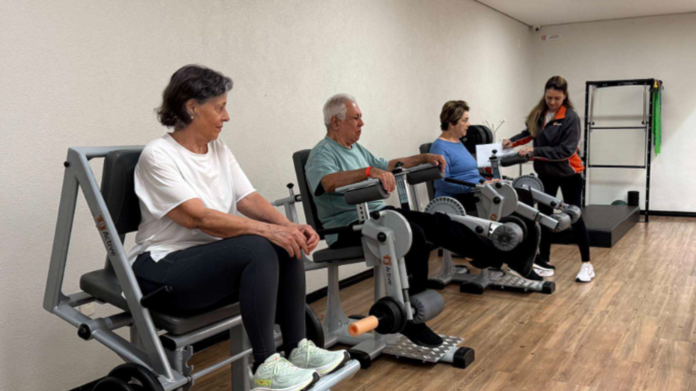 Pessoas idosas em equipamentos de fortalecimento de membros inferiores sob supervisão profissional em Academia Terapêutica.